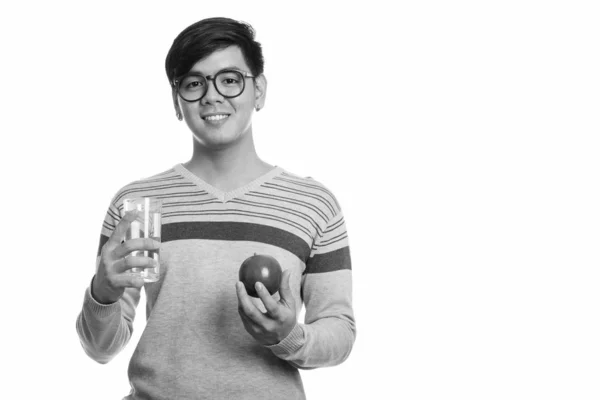 Studioaufnahme eines jungen glücklichen asiatischen Mannes, der lächelnd roten Apfel und ein Glas Wasser in der Hand hält — Stockfoto