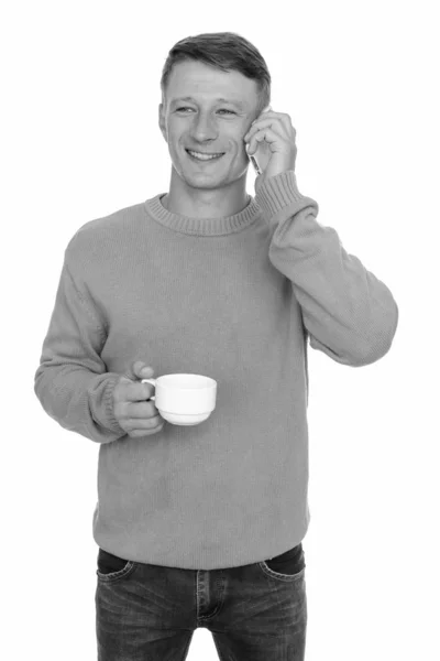 Studioaufnahme eines jungen glücklichen kaukasischen Mannes, der mit dem Handy telefoniert, während er eine Kaffeetasse hält — Stockfoto