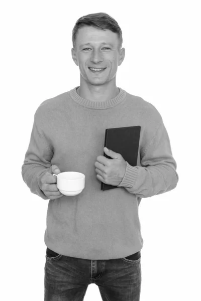 Studio shot of young happy Caucasian man holding book and coffee cup — Stock Photo, Image