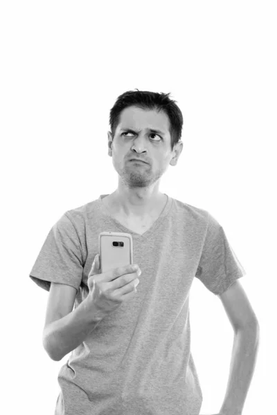 Captura de estudio de un hombre joven sosteniendo el teléfono móvil mientras piensa y mira hacia arriba —  Fotos de Stock