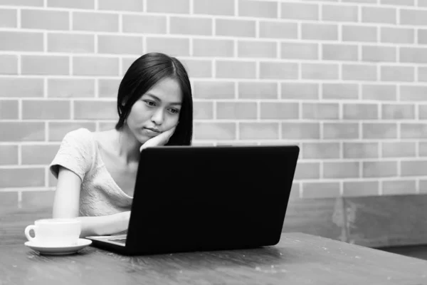 Jeune belle adolescente asiatique reposant menton sur la main tout en utilisant un ordinateur portable avec cappuccino sur une table en bois contre un mur de briques — Photo