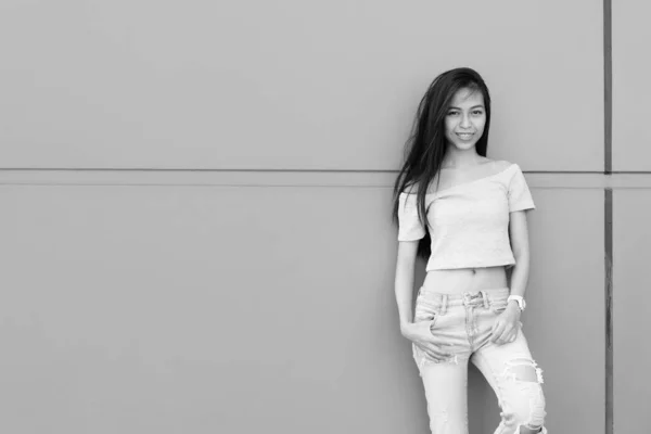 Young happy Asian teenage girl smiling while posing and leaning against concrete wall outdoors — Stock Photo, Image