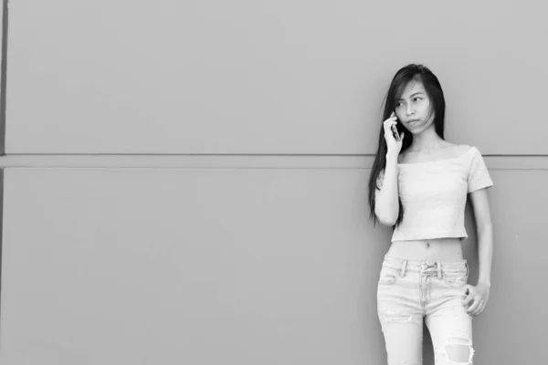 Fiatal gyönyörű ázsiai tinédzser lány beszél a mobiltelefon, miközben áll, és gondolkodás ellen beton fal szabadban — Stock Fotó