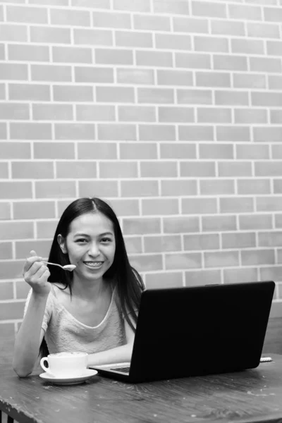 Jeune adolescente asiatique heureuse souriante tout en mangeant la mousse du cappuccino avec ordinateur portable sur une table en bois contre un mur de briques — Photo