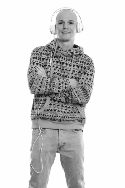 Studio shot of young happy bald man smiling and standing while listening to music with arms crossed — Stock Photo, Image