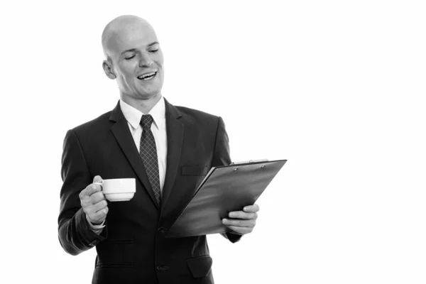 Estudio toma de joven feliz calvo hombre de negocios sonriendo y leyendo en el portapapeles mientras sostiene la taza de café — Foto de Stock