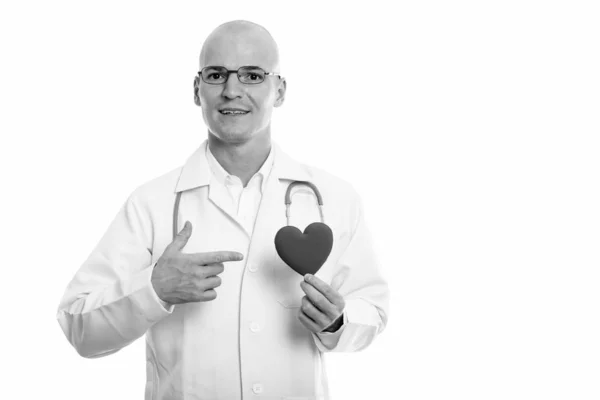 Студийный снимок молодого счастливого лысого мужчины доктор улыбается, держа и указывая на красное сердце — стоковое фото
