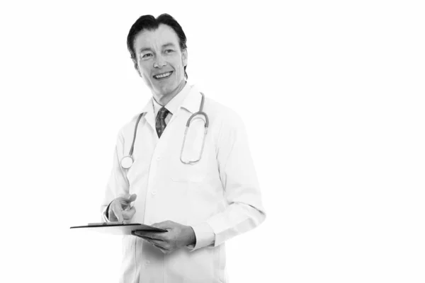 Studio shot de mature homme heureux médecin souriant et pensant tout en tenant presse-papiers — Photo