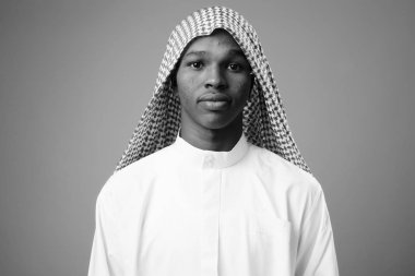 Studio shot of young African man wearing traditional Muslim clothes against gray background in black and white