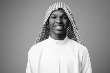 Studio shot of young African man wearing traditional Muslim clothes against gray background in black and white