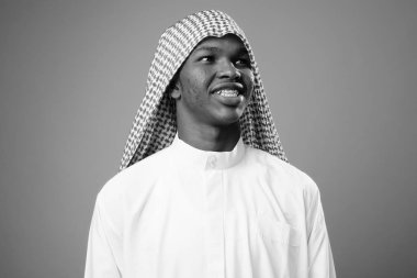 Studio shot of young African man wearing traditional Muslim clothes against gray background in black and white