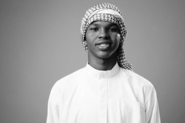 Studio shot of young African man wearing traditional Muslim clothes against gray background in black and white