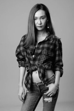 Studio shot of young beautiful Asian hipster woman against gray background in black and white
