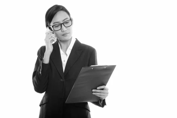 Studio shot van jonge Aziatische zakenvrouw praten op mobiele telefoon terwijl op zoek naar klembord — Stockfoto