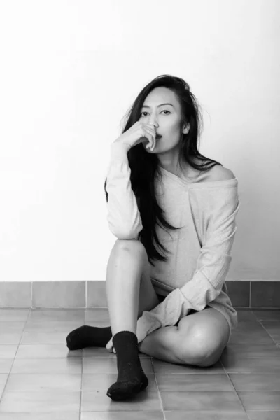 Studio plan de jeune belle femme asiatique assise sur le sol et posant avec la main sur la bouche — Photo