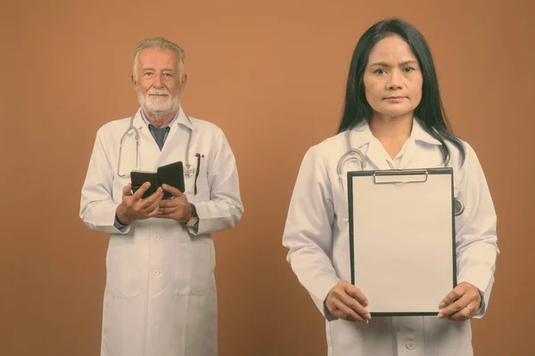 Studio shot of senior man doctor and mature Asian woman doctor together against brown background