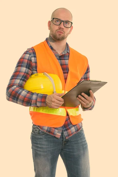 Calvo musculoso trabajador de la construcción sosteniendo casco de seguridad y portapapeles mientras piensa —  Fotos de Stock
