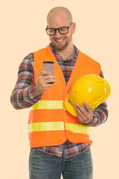 Щасливий лисий м'язистий чоловік будівельник посміхається, тримаючи шолом безпеки і використовуючи мобільний телефон — стокове фото