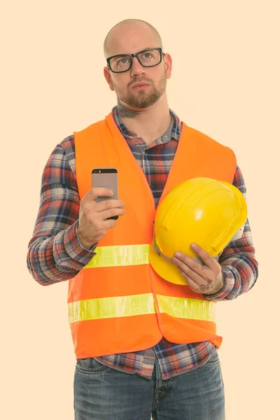 Pensativo careca homem muscular trabalhador da construção segurando capacete de segurança e telefone celular — Fotografia de Stock