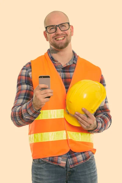 Pensativo feliz careca homem muscular trabalhador da construção sorrindo enquanto segurando capacete de segurança e telefone celular — Fotografia de Stock