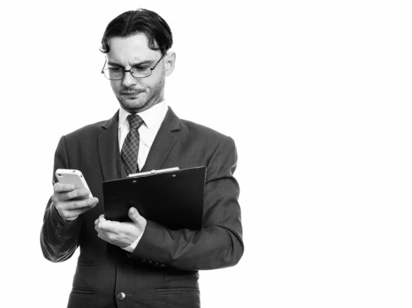 Captura de estudio de un joven hombre de negocios usando el teléfono móvil mientras sostiene el portapapeles —  Fotos de Stock