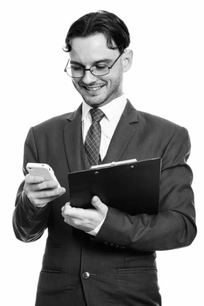 Studio shot of young happy businessman smiling while using mobile phone and holding clipboard — Stock Photo, Image