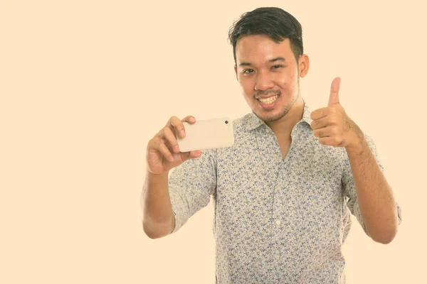 Young happy Asian man smiling while taking picture with mobile phone and giving thumb up — Stock Photo, Image