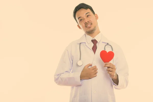 Στούντιο πλάνο του νεαρού Ασιάτη γιατρό κρατώντας κόκκινη καρδιά, ενώ σκέφτεται — Φωτογραφία Αρχείου