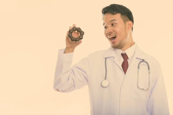 젊고 행복 한 아시아인 의사가 도넛을 들고 보면서 웃는 모습이 담긴 스튜디오 사진 — 스톡 사진