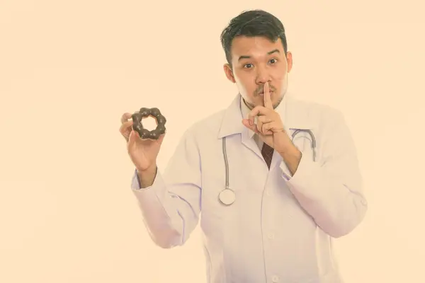 입술에 도넛을 손가락으로 쥐고 있는 젊은 아시아 남자 의사의 스튜디오 촬영 — 스톡 사진