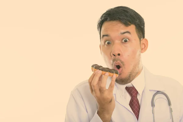Studio strzał młody Azji człowiek lekarz jedzenie pączka podczas patrząc zszokowany — Zdjęcie stockowe