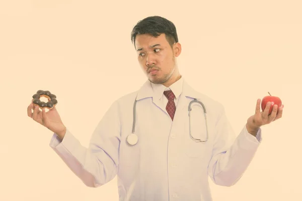 Studio shot of young Asian man doctor looking at donut and holding red apple — Stock Photo, Image