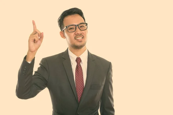 Studio shot van jong gelukkig aziatisch zakenman glimlachen terwijl wijzend vinger omhoog — Stockfoto