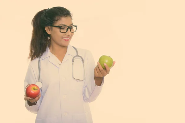Estudio disparo de joven feliz médico mujer persa sonriendo mientras sostiene manzana roja y mirando manzana verde aislado sobre fondo blanco — Foto de Stock