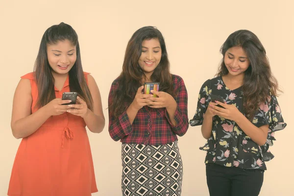 Estúdio tiro de três felizes jovens persas mulheres amigas sorrindo ao usar telefones celulares juntos contra fundo branco — Fotografia de Stock