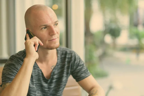 Young handsome bald man thinking while talking on mobile phone inside restaurant with glass windows