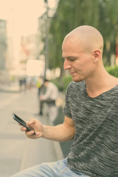 Profile view of young handsome bald man using mobile phone while sitting outdoors — Stock Photo, Image