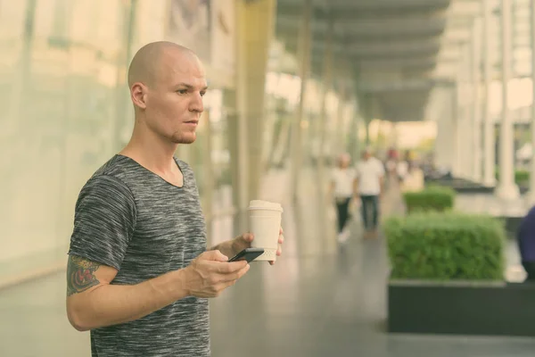 Young handsome bald man holding paper coffee cup and mobile phone while thinking outside of the mall in Bangkok Thailand — Stock Photo, Image