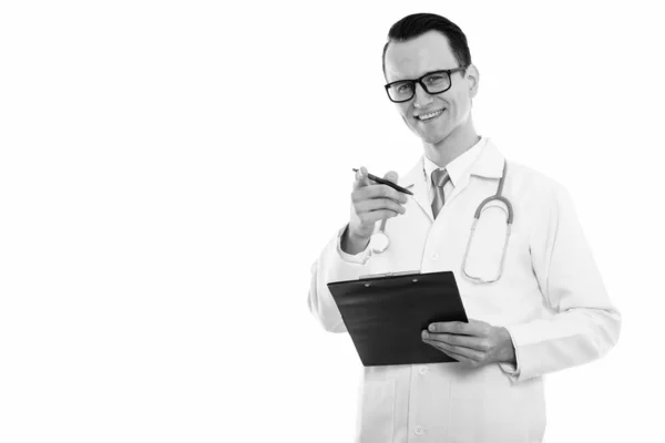 Studio shot of young happy man doctor smiling while holding clipboard and pen while pointing at camera — Stock Photo, Image