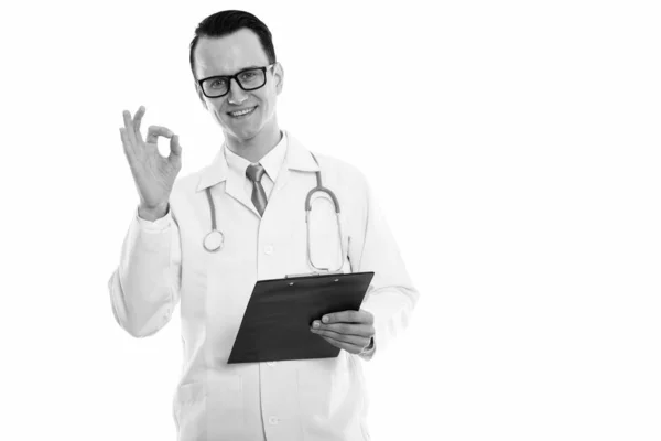 Studio shot of young happy man doctor smiling while holding clipboard and giving ok sign — Stock Photo, Image