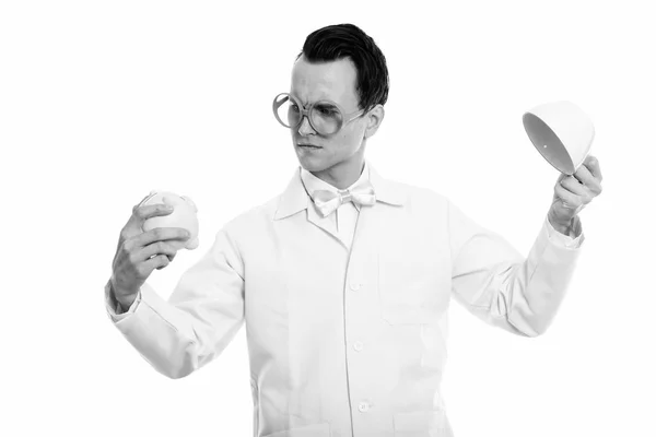 Studio shot of young crazy man doctor looking at piggy bank while holding empty coffee cup upside down — Stock Photo, Image