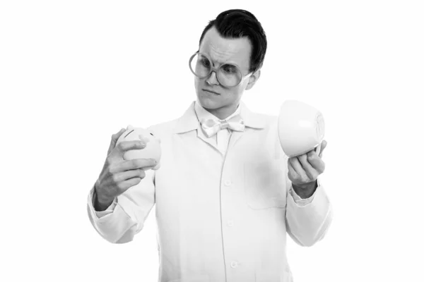 Studio shot of young crazy man doctor looking at piggy bank while holding empty coffee cup — Stock Photo, Image
