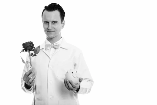 Studio shot of young man doctor holding red rose and piggy bank — Stock Photo, Image