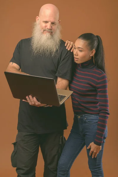 Mature bearded bald man with young Asian transgender woman against brown background