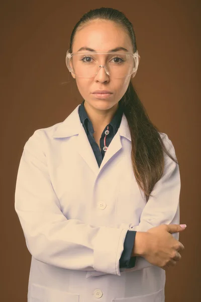 Ung vacker kvinna läkare bär skyddsglasögon mot brun bakgrund — Stockfoto