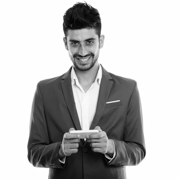 Studio shot of young happy Persian businessman smiling while using mobile phone and looking at camera — 스톡 사진
