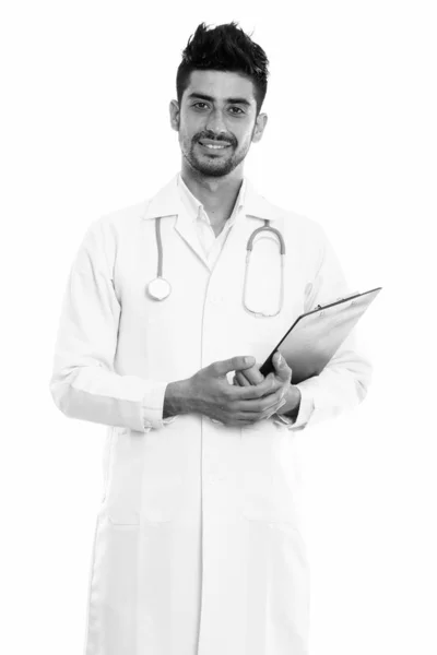 Estudio disparo de joven feliz hombre persa médico sonriendo y de pie mientras sostiene el portapapeles —  Fotos de Stock