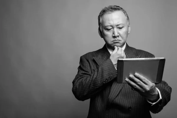Studio shot of mature Asian businessman using digital tablet against gray background in black and white