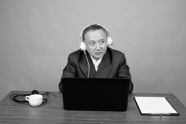 Gammal asiatisk affärsman sitter med träbord mot grå bakgrund — Stockfoto