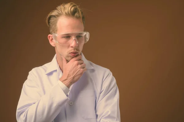 Mladý pohledný muž lékař nosí ochranné brýle proti hnědému pozadí — Stock fotografie